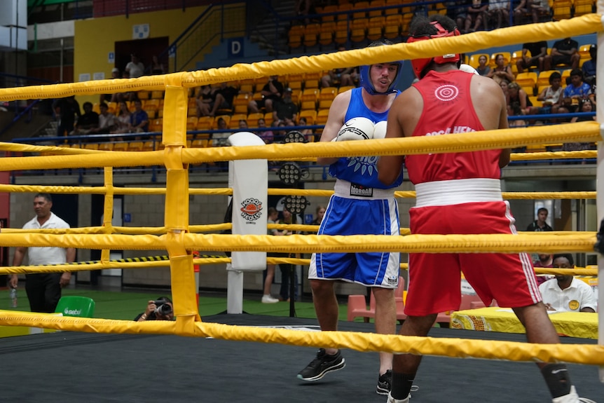 Two boxers wearing headgear compete inside a ring, one is wearing blue and the other is dressed in red.