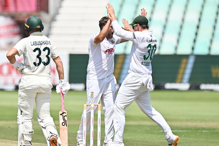 A cricket player in white holds his bat while players celebrate around him