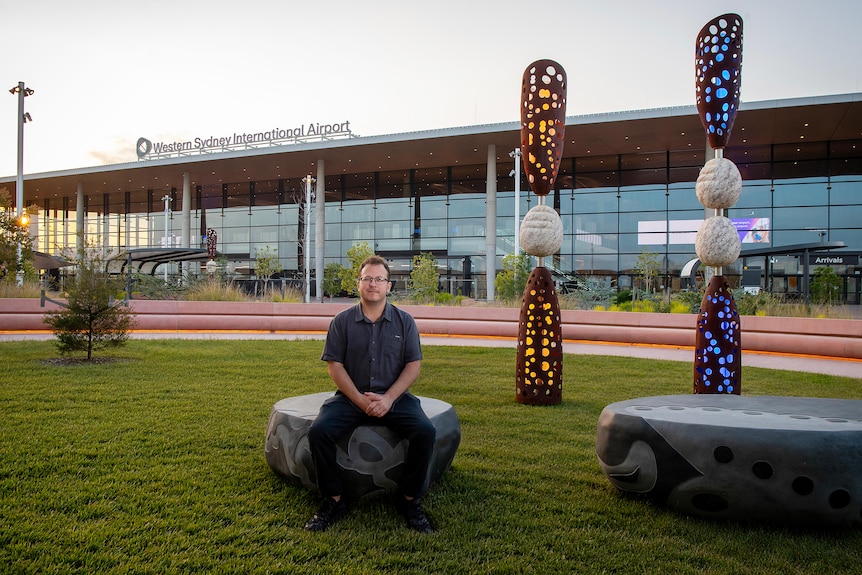 A man sitting outside an airport terminal called Western Sydney International, next to Indigenous sculture artwork, in the AM.