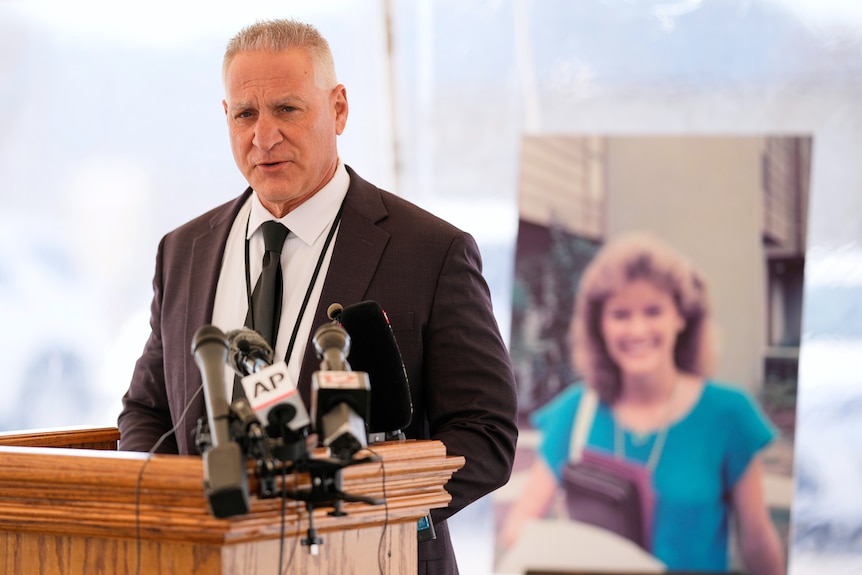 A man talks behind a microphone, with a photo of a young woman behind him.
