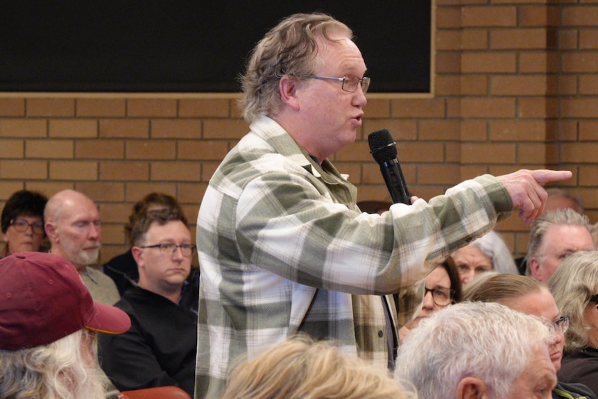 A midddle-aged man points as he stands and speaks into a microphone among a crowd in a community hall.