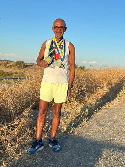 An older athlete proudly displaying multiple medals while standing on a gravel path with dry grass in the background. An older athlete proudly displaying multiple medals while standing on a gravel path with dry grass in the background.