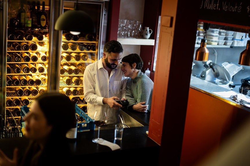 A woman puts her head on a man's shoulder with a restaurant and bar behind them