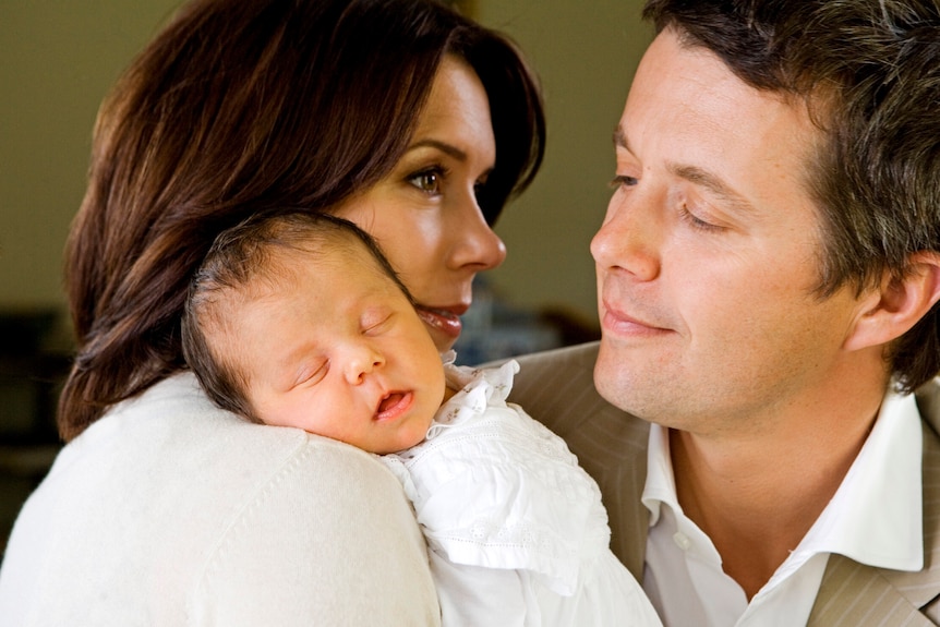A baby lies on a woman's shoulder and a man looks on