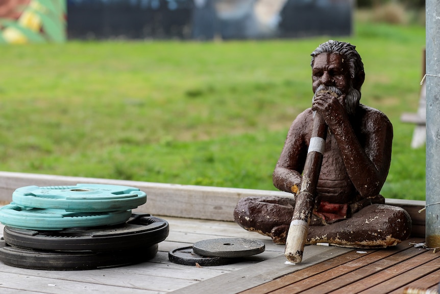 A small statute fo an Aboriginal man playing the didgeridoo next to some weights outside in a yard