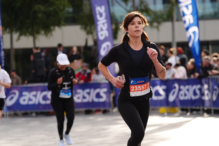 A young woman dressed in a black t-shirt and black tights holds her phone as she runs.