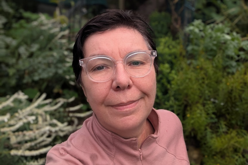 A woman with glasses, short hair and a rose pullover in an outdoor area.