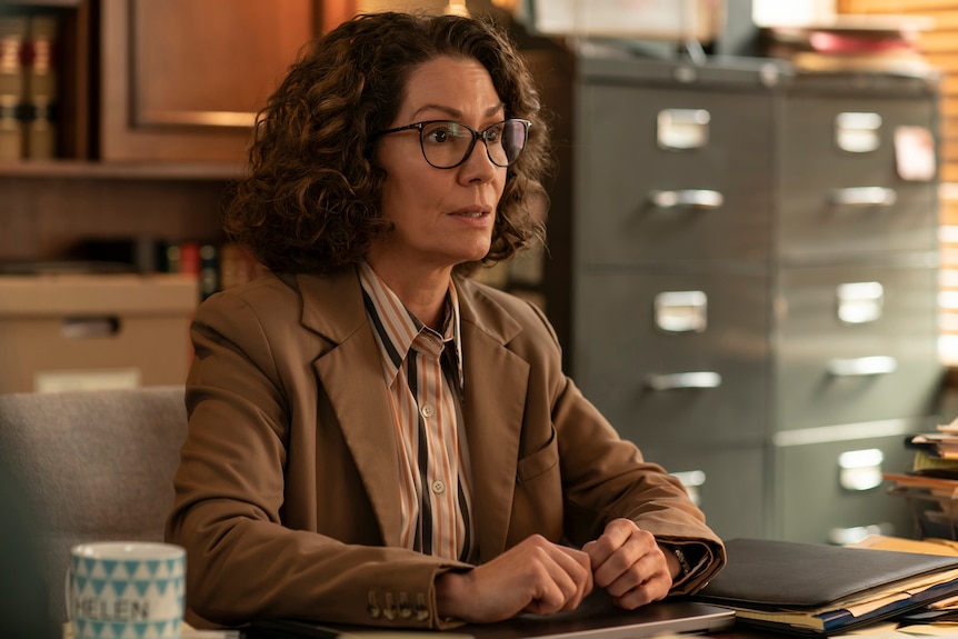 A TV still of Kitty Flanagan sitting at a desk, wearing glasses and a loose brown suit. She has a bemused expression.