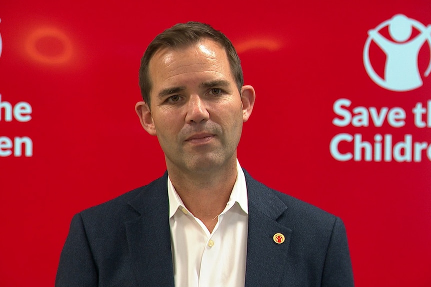A man speaking to media in front of a bright red backdrop