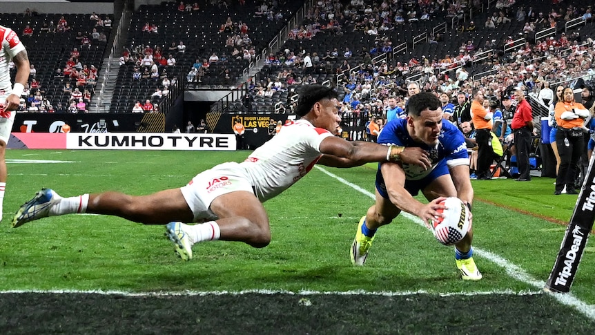 A Bulldogs NRL player dives over in the corner for a try past a Dragons player in Las Vegas, with empty seats behind them. 
