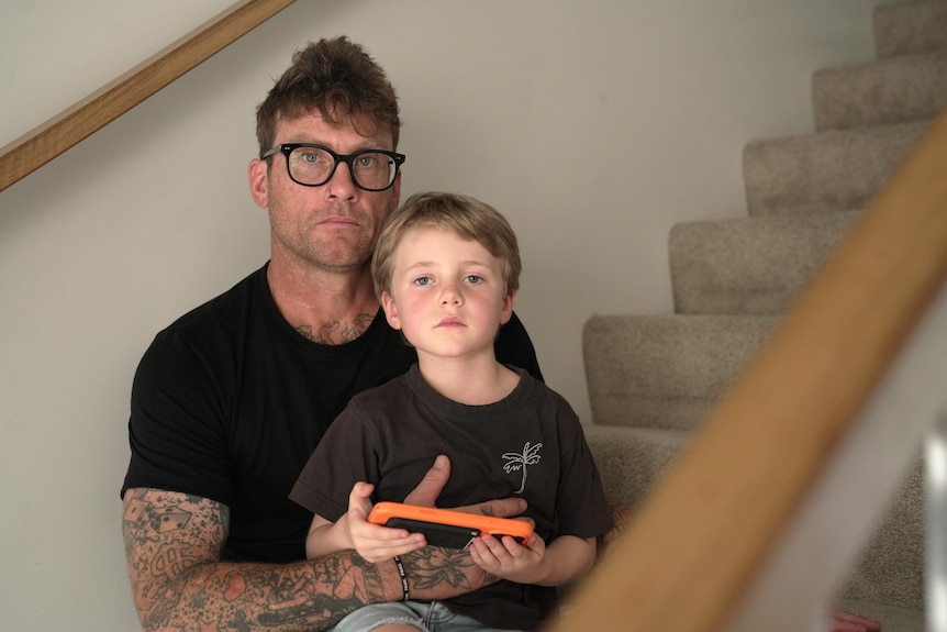 A man sits on a staircase with his son on his knee. Both look into camera with serious expressions.