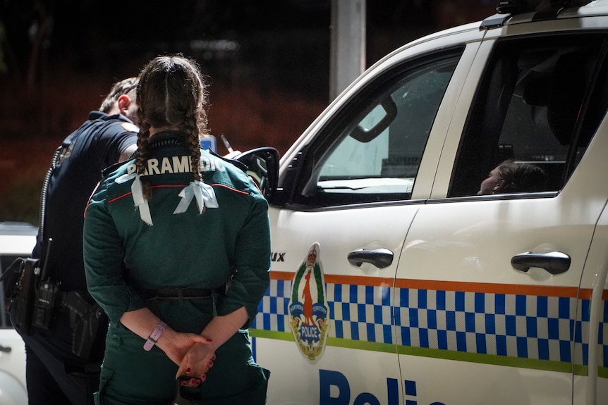 A woman viewed from the back with her hair in two plaits wearing a paramedic uniform stands beside a police car and an officer