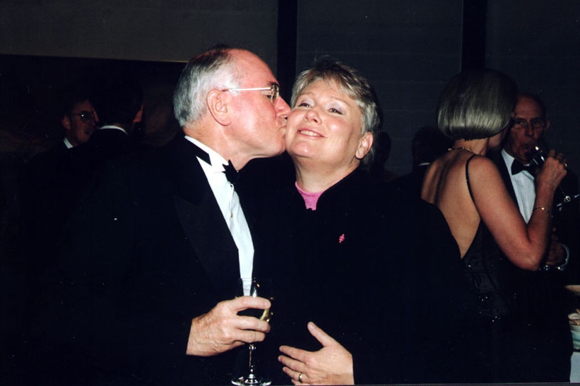 A man in a black suit and holding a glass of wine, kisses a woman on the cheek.