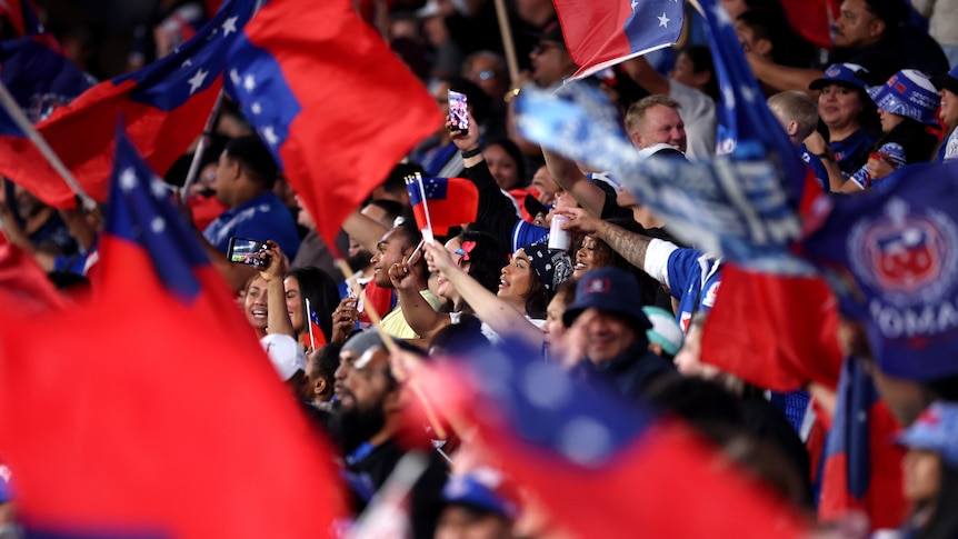 A packed crowd of Samoan rugby league fans cheer and wave flags during a game.