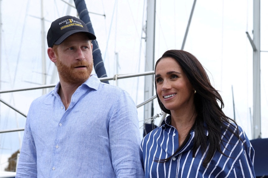 A man and woman (Prince Harry and Meghan Markle) stand with boat masts in the background.