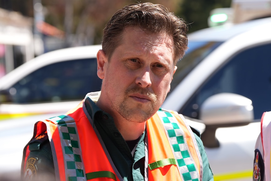 A man listens during a press conference outside, wearing hi-vis.