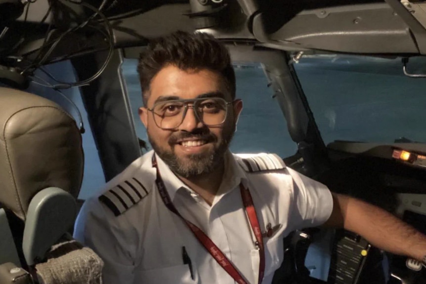 An Indian man sits in the cockpit of a big plane, with a crisp white uniform and a big smile.