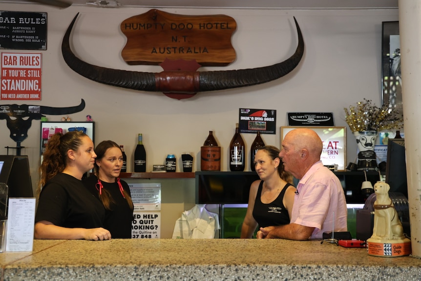 Three women in black shirts and man in a pink shirt stand chatting behind a bar in a pub.