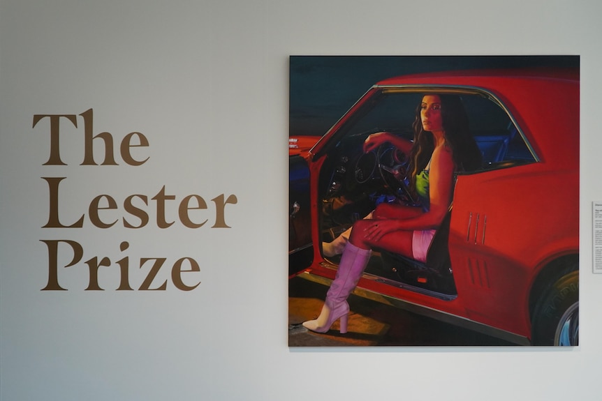 A painting of a woman driving a car is hung on a wall next to the words 'The Lester Prize'.