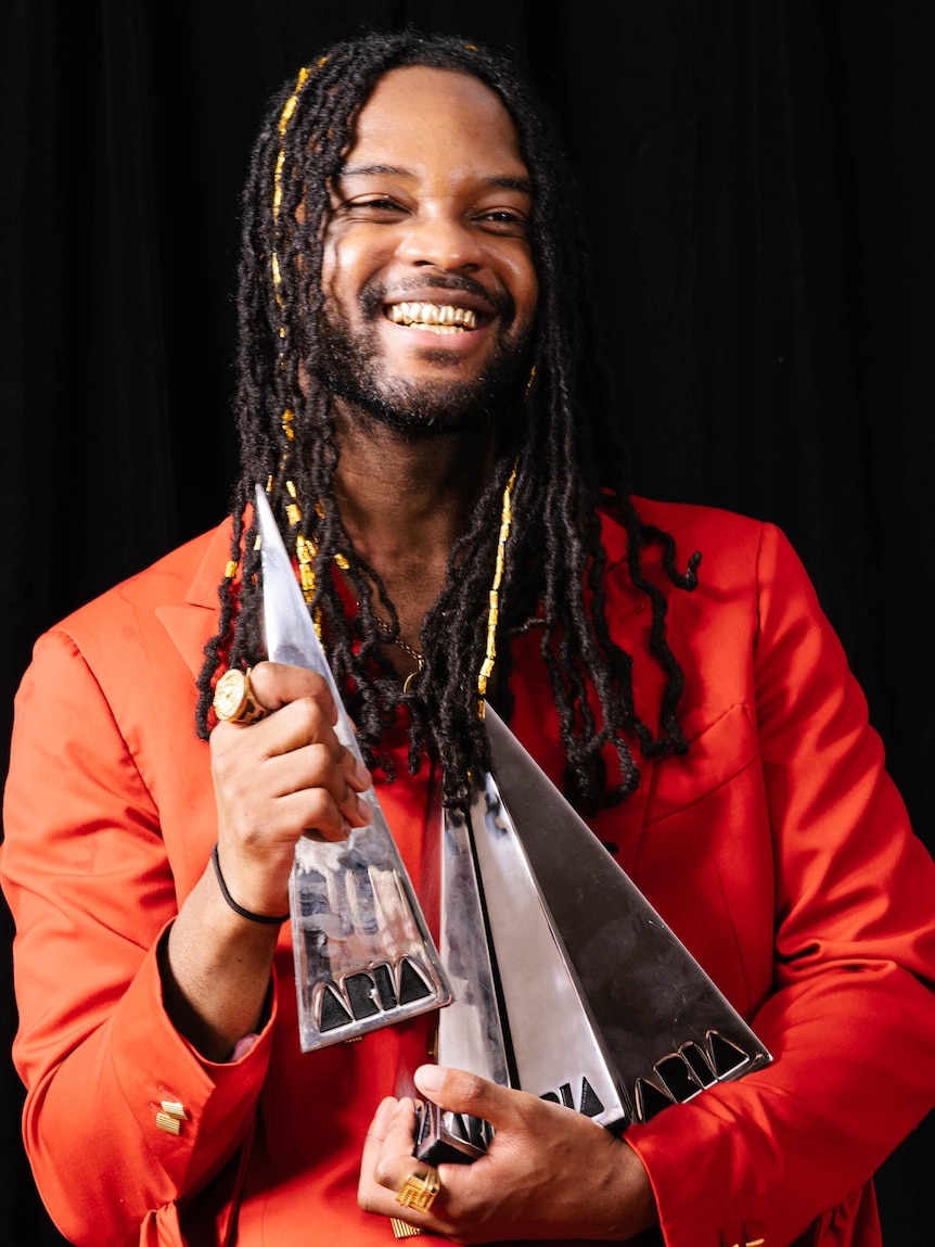 Smiling Ghanian-Australian man iin red jacket clutches three pointy silver award trophies