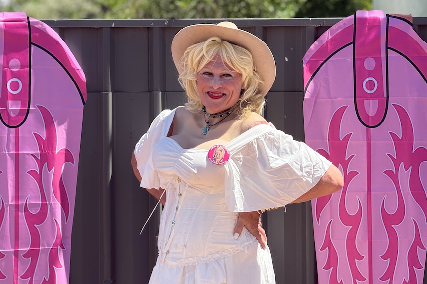 A person wearing a white summer dress and blonde wig with a broad-brimmed hat poses against a black fence.