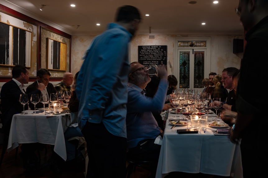 A man swigs the last of a glass of wine, surrounded by full restaurant tables, as a wine waiter looks on. 