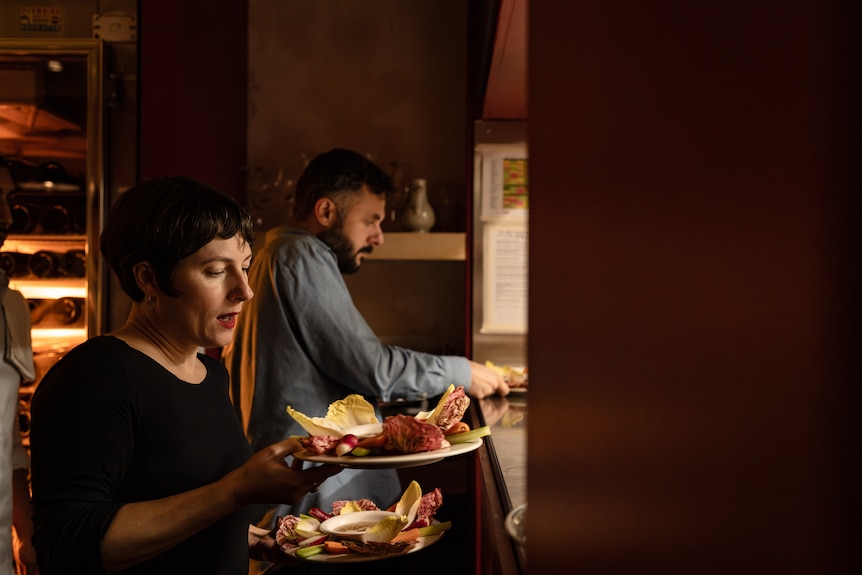 Two people carry plates of food with glow of restaurant kitchen on their faces