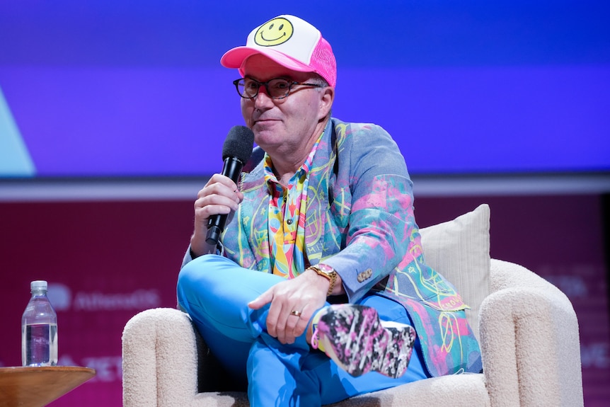 Dan Ives speaking on a couch in a brightly coloured jacket and shirt and a cap with a smiley face on it.