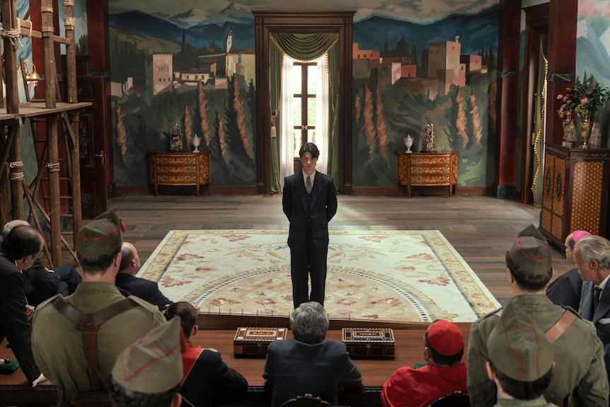 A young man in a black suit stands in front of a group of older men in a baroque building