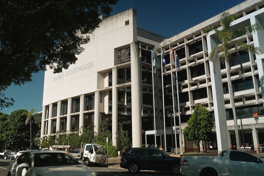 Looking across the road at a five story concrete and glass building. In front of it are flagpoles and palm trees.
