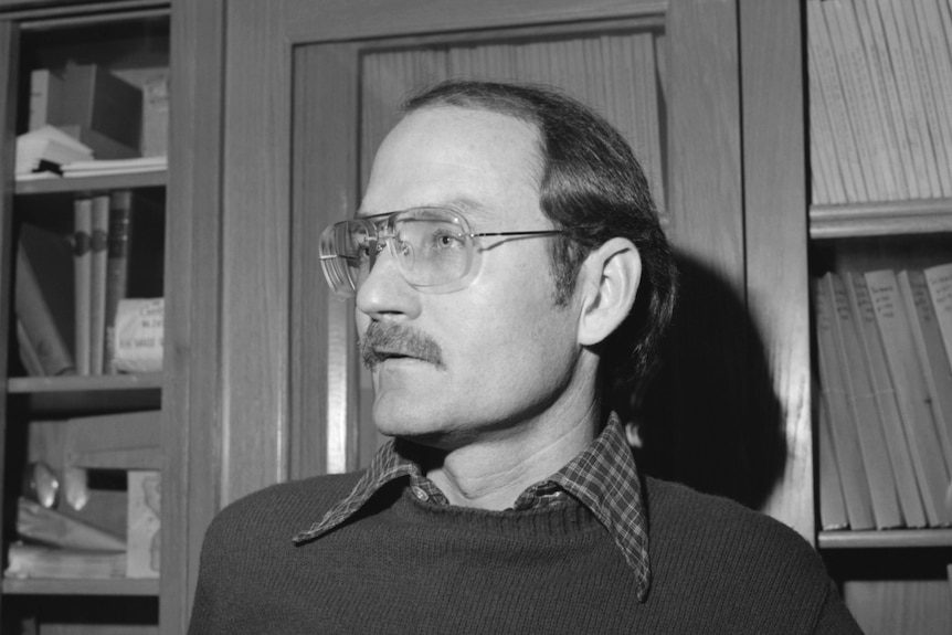 Black and white photograph of a man with a mustche and glasses. Image is shoulder up, man is looking to the left of the frame.