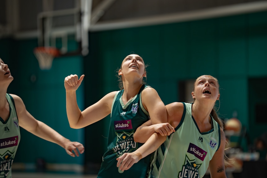 Two Jewels players dressed in green compete for the ball in an indoor basketball court with green walls