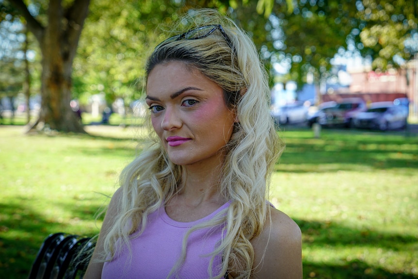 A woman with bleached blonde hair wearing a pink tank top sitting in a park.