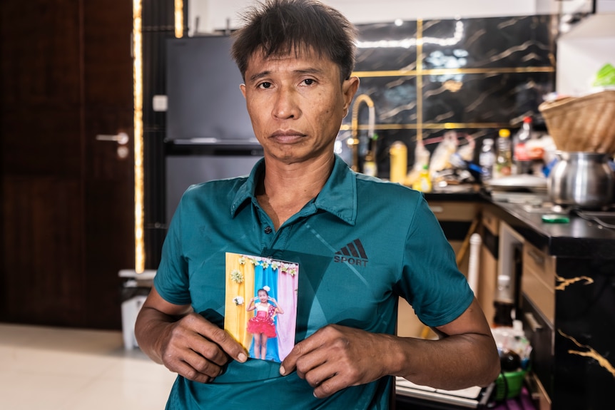 A close up of a Thai man wearing a green shirt sitting in his kitchen while holding a photo of his daughter.