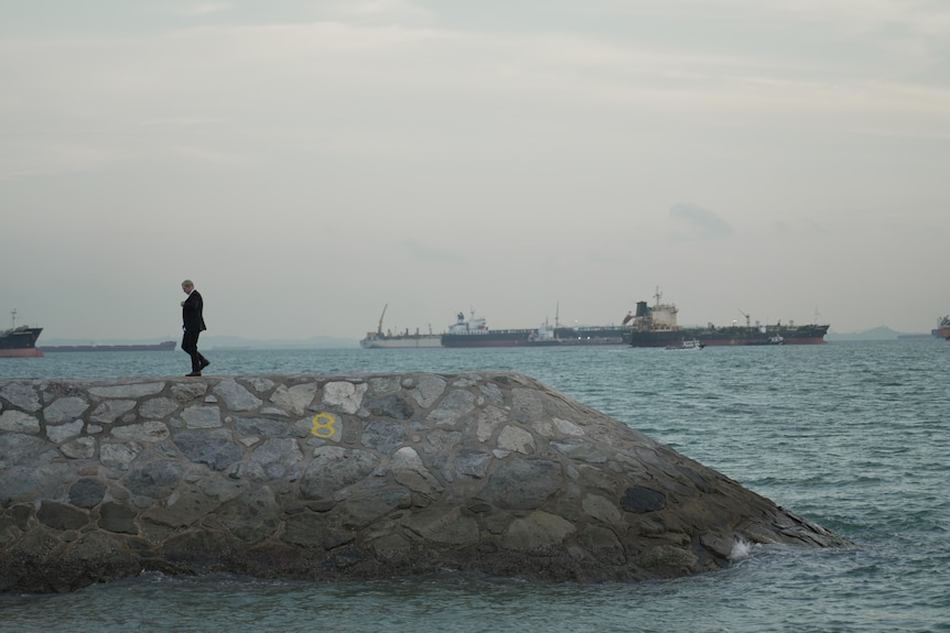 A stone jetty with a man walking on it, with large ships in the background.