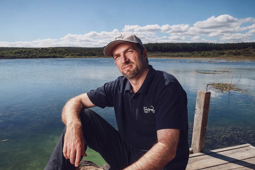 A man wearing a baseball cap and navy blue polo shirt sits on a wetland pier, arm on his knee.