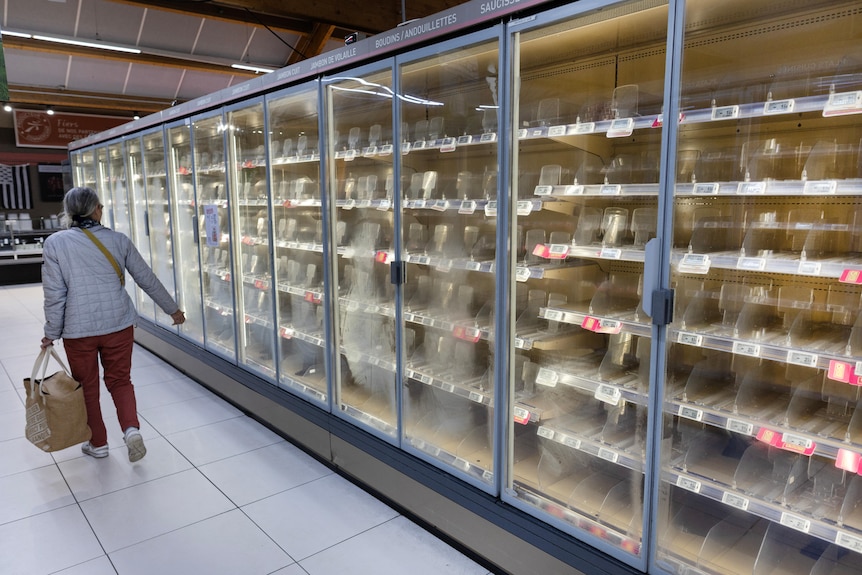 A woman walks past an empty freezer section in a supermarket.