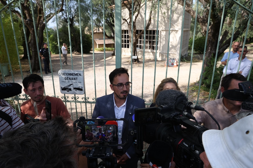 a man flanked by reporters tands in front of a closed gate