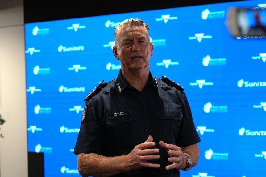 A man in a Victoria Police uniform gestures as he speaks in front of a blue background.
