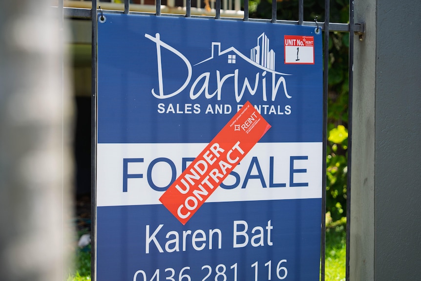 A blue 'for sale' sign on a fence outside a block of units in Darwin, with a red 'under contract' sticker plastered over it.