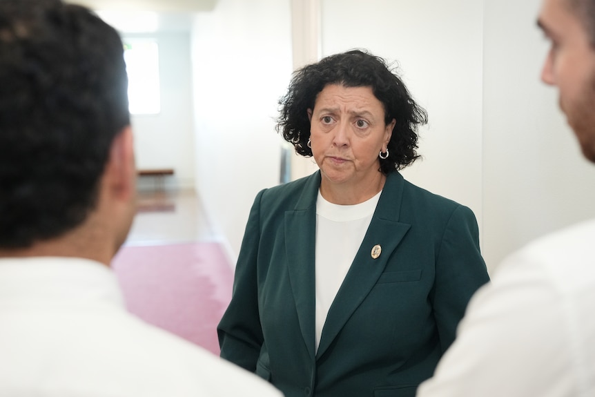 A dark-haired woman (Monique Ryan) in a suit jacket looks concerned as she talks to doctors.