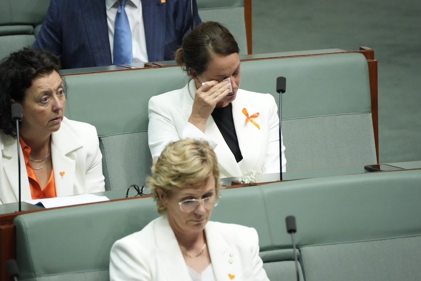 Sophie Scamps wipes her eyes while wearing white blazer and orange ribbon in Parliament.