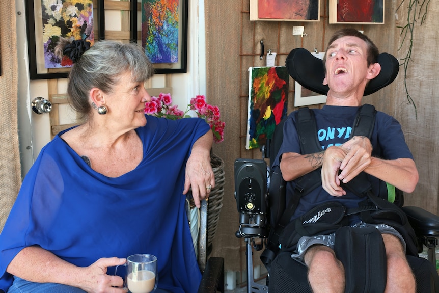 Wendy and Colin Hanssen sit side by side in front of artwork on a wall.