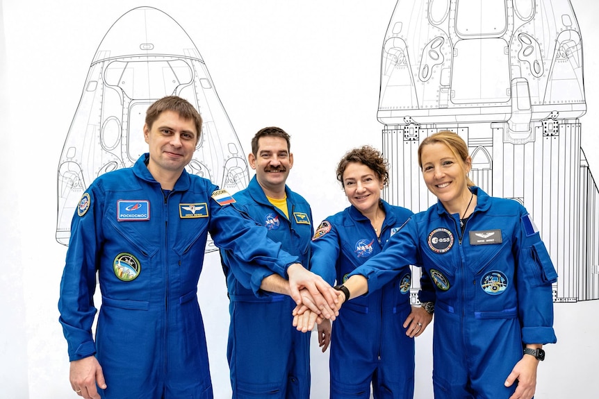 Male and female astronauts in blue space jumpsuits, smiling while placing their hands in a single pile.