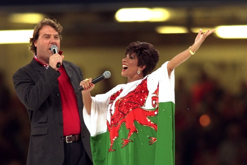 Bryn Terfel and Shirley Bassey perform on stage, Bassey in a kaftan dress of the Welsh flag, before a stadium audience.
