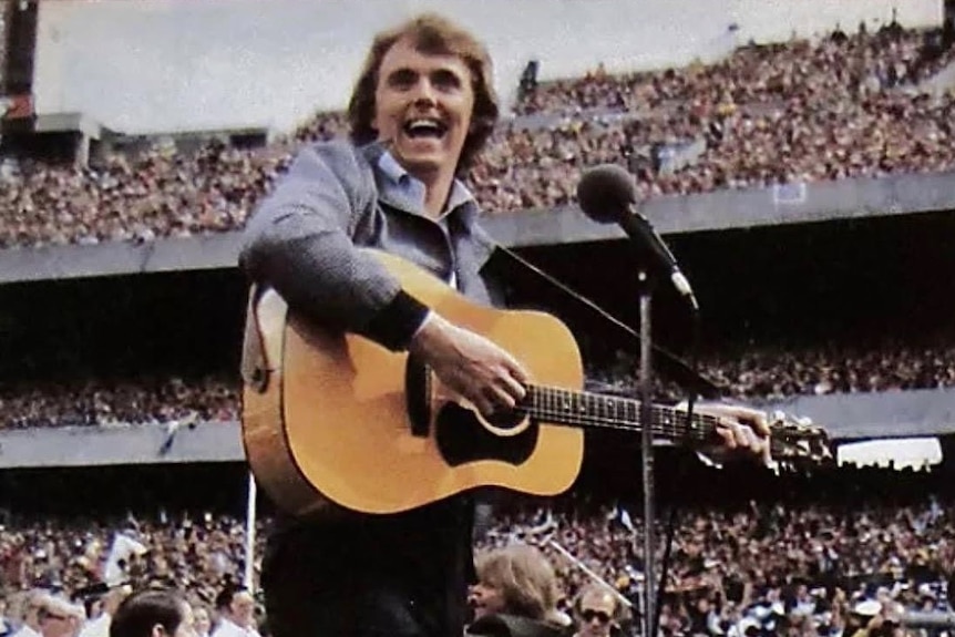 Singer-songwriter Mike Brady performing at the 1979 VFL Grand Final, holding a guitar and singing to a crowd.