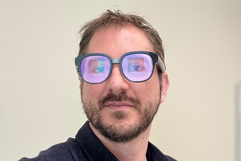 A bearded man with short hair and wearing dark-rimmed glasses that have a purplish glow.