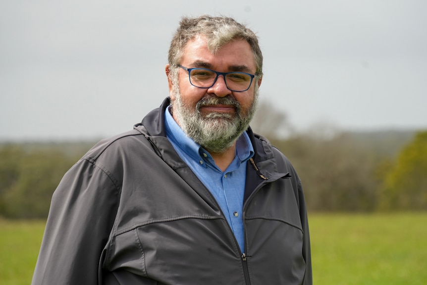 Damein Bell standing and smiling in front of the camera, a green landscape behind him.