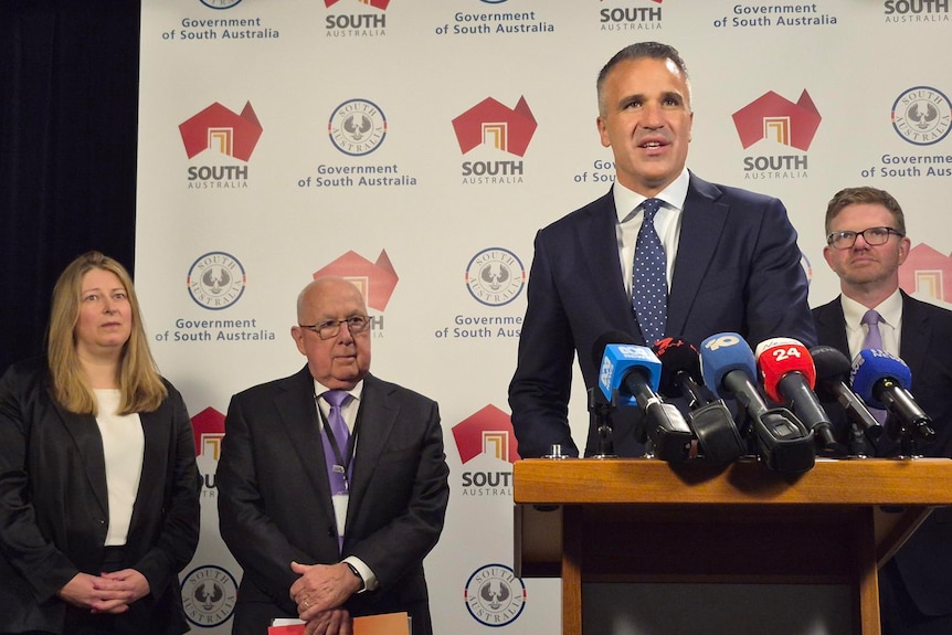 Peter malinauskas stands in front of microphones in front of Iain Ross, Kate Pounder and Chris Picton