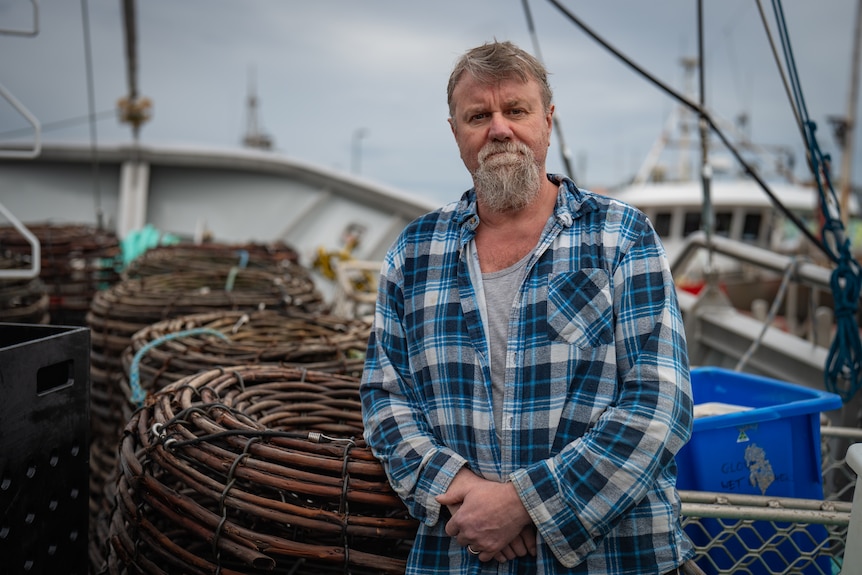 A man in a blue plaid shirt and a beard leans against a stack of lobsters pots